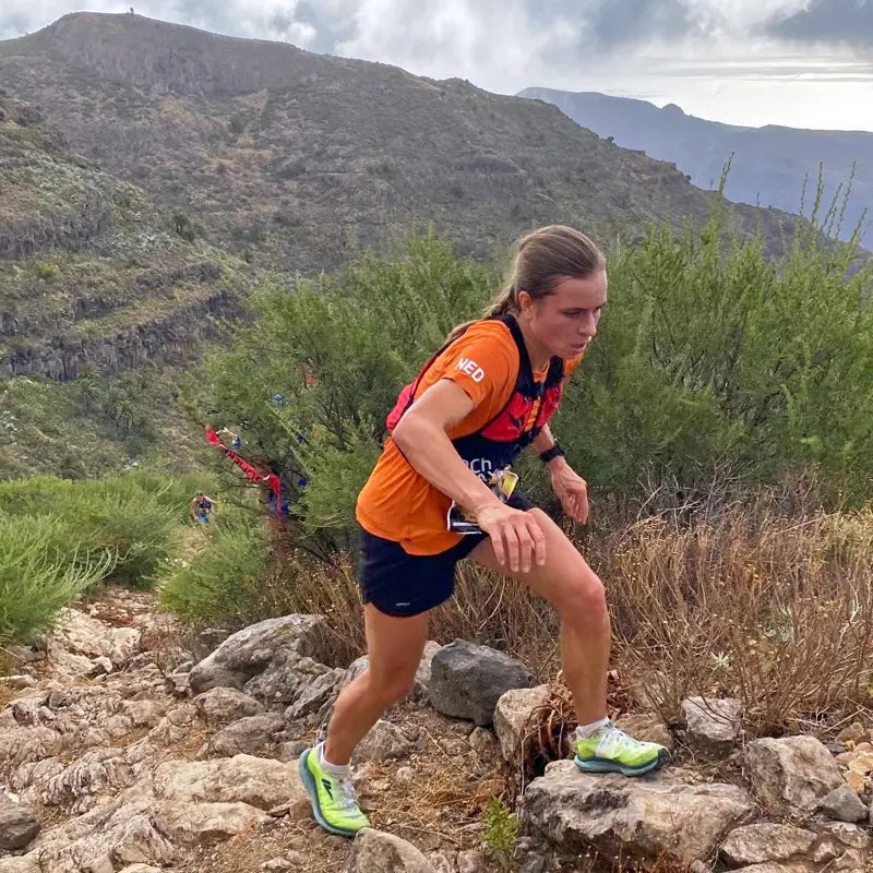 Een persoon in een oranje shirt die over een rotsachtig, bergachtig pad hardloopt, met groene heuvels op de achtergrond.
